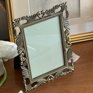 Peut inclure: Un cadre photo rectangulaire avec une finition en laiton antique, orné de volutes et de détails en forme de feuilles sur les bords. Le cadre présente une bordure intérieure perlée et deux petits accents en forme de cœur en haut et en bas.