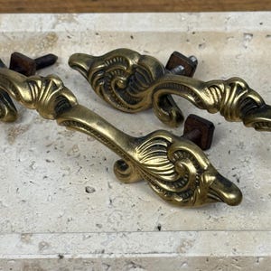 May include: Close-up of four antique brass drawer pulls with ornate, swirling designs. Each pull is attached to a wooden block with a screw. The pulls are arranged on a light-coloured, textured surface.