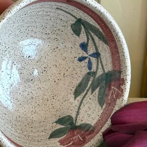 May include: A speckled ceramic bowl with a floral design. The bowl has a red rim and is decorated with green leaves, blue flowers, and red flower buds. The bowl is a light beige colour.