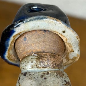 May include: A ceramic figurine of a person wearing a dark blue hat with a light blue and tan brim. The figurine has a tan head and a speckled, light grey body. The collar is a speckled tan colour.