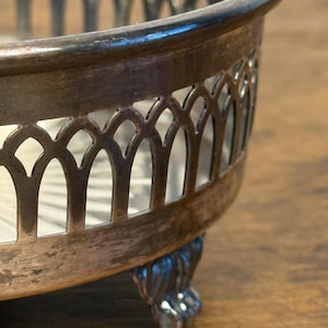 Peut inclure: Gros plan sur un plat de service en métal argenté avec un motif ajouré décoratif. Le plat a un bord arrondi et repose sur trois pieds ornés en forme de griffes. Le fond est une surface en bois.