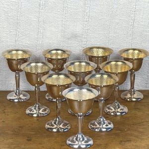 May include: Ten silver goblets with gold-coloured interiors are displayed. The goblets have a hammered texture inside the bowls. The stems and bases are silver, and the goblets are arranged on a wooden surface.