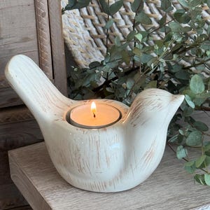 May include: A bird-shaped candle holder made of off-white ceramic with a distressed, wood-grain texture finish. The centre of the bird features a circular opening holding a small, lit tea light candle with a visible orange flame and metal wick.