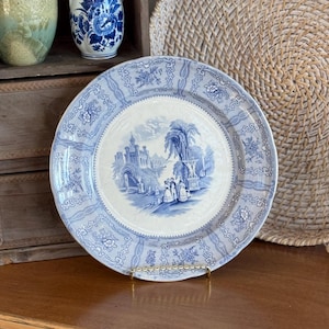 May include: A round ceramic plate featuring a blue and white transferware pattern. The center depicts a landscape with a castle, trees, and figures. The wide rim is decorated with repeating floral motifs and geometric borders in blue on white.