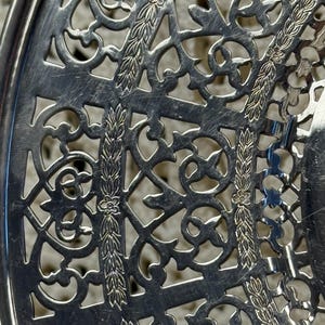 May include: Close-up of a silver metal decorative tray with an openwork design. The tray features intricate patterns of swirling lines, floral accents, and geometric shapes. The metal has a polished, reflective surface.