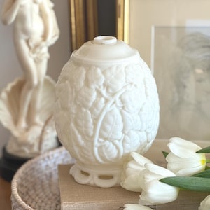 Peut inclure: Un pot décoratif blanc en forme d'œuf avec un couvercle, orné d'un motif de feuilles en relief. Le pot repose sur une petite base et est entouré de tulipes blanches. Une statue et des œuvres encadrées sont en arrière-plan.