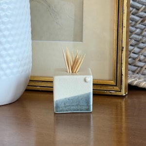 May include: A small, cube-shaped ceramic toothpick holder featuring a two-tone design with a cream-coloured top half and a speckled, slate-blue bottom half. A small, raised heart detail is visible on the top right corner, filled with wooden toothpicks.