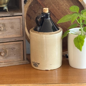 May include: A cylindrical stoneware jug with a beige body and a dark brown glazed top section. The jug features a single handle, a cork stopper, and a small black stamped crown logo near the base. The vessel has a smooth, matte finish on the lower portion and a glossy finish on the upper neck.