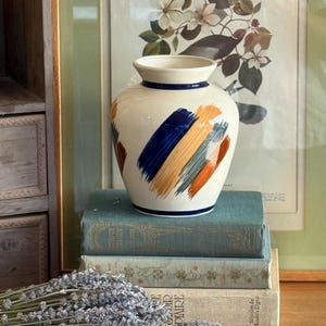May include: A cream-coloured ceramic vase with a wide, rounded body and a narrow neck. The exterior features bold, diagonal brushstroke patterns in shades of navy blue, golden yellow, and burnt orange, with thin navy blue bands at the rim and base.