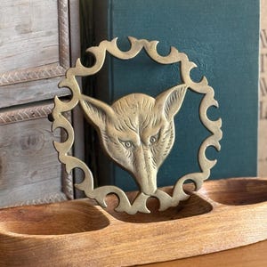 May include: A circular brass trivet featuring a detailed fox head at the centre, surrounded by a decorative scalloped border. The brass piece is positioned upright within a long, three-sectioned wooden tray with a natural light brown finish.