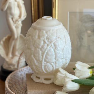 May include: A white, egg-shaped decorative jar with a lid, featuring a detailed leaf pattern. The jar sits on a decorative base. White tulips and a statue are in the background, suggesting a vintage or antique theme.