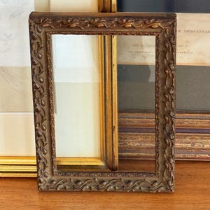 May include: A rectangular, ornate, antique picture frame with a dark brown finish. The frame features detailed floral and scrollwork carvings. Other frames are visible in the background, suggesting a collection or display of art.