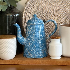 Pode incluir: Uma cafeteira esmaltada azul e branca com tampa e alça. A cafeteira tem um padrão salpicado e é exibida com outros itens de cozinha, incluindo um pote branco com tampa de madeira e uma bandeja tecida.