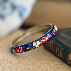 May include: A gold bangle with a dark blue enamel band, adorned with red, white, and pink floral patterns. The bracelet is displayed on a tan surface, next to a book. The bracelet's design is intricate.