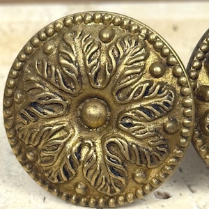 May include: Close-up of two antique brass cabinet knobs. Each knob is round with a detailed floral design in the centre, surrounded by a beaded edge. The knobs have a warm, aged patina, suggesting they are vintage or vintage-inspired.