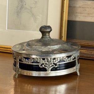 May include: An antique silver-plated trinket box with a black glass base and ornate floral detailing. The lid features a textured design and a small, decorative knob. The box is set on small, curved feet.