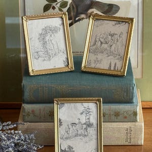 Op de afbeelding: Drie rechthoekige fotolijsten van goudkleurig metaal met kralenranden. Elke lijst bevat een zwart-wit print in toile-stijl met pastorale scènes met figuren, paarden en bomen. De lijsten zijn in een groep van drie gerangschikt.