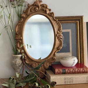 Peut inclure: Un miroir ovale doré orné d'un cadre détaillé. Le miroir est présenté sur une surface en bois, à côté d'un petit vase avec des fleurs, des livres et une image encadrée. La scène évoque une esthétique vintage et antique.