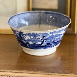 Peut inclure: Un bol en céramique bleu et blanc avec une scène de paysage détaillée. Le bol présente une base à pied et un bord à motifs. Le design comprend des personnages, des bâtiments et des arbres, créant une esthétique classique et vintage. Le bol est présenté sur une surface en bois.