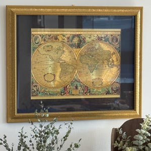 Op de afbeelding: Een ingelijste antieke wereldkaart met de titel NOVA TOTIVS TERRARVM ORBIS GEOGRAPHICA AC HYDROGRAPHICA TABVLA, met twee gouden cirkelvormige hemisferen, decoratieve illustraties en een donkere passe-partout in een getextureerde gouden lijst.