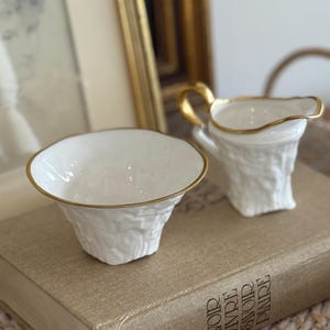 Peut inclure: Un ensemble de crème et de sucrier en porcelaine blanche avec des bordures dorées. La crémière a une anse et un bec dorés. Le sucrier est rond avec un bord doré. Les deux pièces ont un motif texturé en forme d'écorce. L'ensemble est présenté sur un livre.