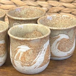 May include: A collection of handmade ceramic cups with a speckled beige and brown glaze. Each cup has a white, abstract design. The cups are cylindrical with a slightly flared rim, ideal for serving drinks.