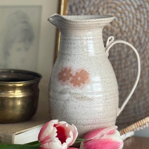 May include: A speckled white ceramic jug with a handle and a spout, decorated with two pink flower designs. The jug is next to pink and white tulips and a brass pot. A book and a framed picture are in the background.