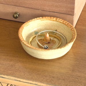 Könnte beinhalten: Eine kleine, runde Schmuckschale aus Keramik mit einem erhöhten Ringhalter in der Mitte. Die Schale ist beige mit einem strukturierten Rand und enthält mehrere goldene und silberne Armbänder. Im Hintergrund befindet sich eine Holzkiste.