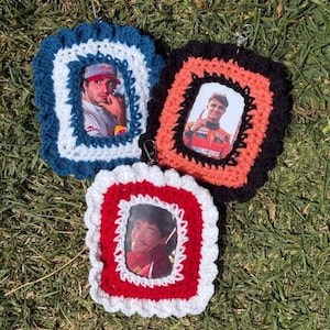 May include: Three handmade crochet picture frames in blue, black, and red, each with a different photo of a person. The frames have a scalloped edge and a small metal clasp. The frames are lying on a bed of green grass.