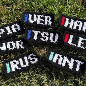 May include: A collection of black crochet patches with white lettering spelling out various names, each with a colored stripe. The patches are arranged on a bed of green grass. The names include VER, HAM, LEC, TSU, NOR, RUS, and IANT.