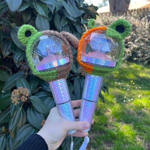 Pode incluir: Duas varinhas de luz com globos transparentes e facetados e cabos iridescentes são seguradas em uma mão. Cada varinha é adornada com um chapéu de orelha de animal de crochê, um verde e marrom, o outro laranja e verde. Um cordão branco pende de cada cabo.