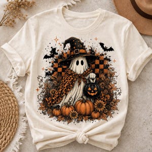 May include: A cream-coloured t-shirt featuring a graphic of a ghost wearing a black witch hat and a leopard print cape. The ghost holds a jack-o'-lantern bucket, surrounded by orange pumpkins, sunflowers, and black bats against a checkered background.