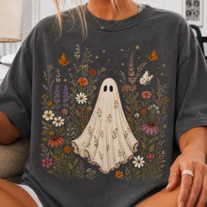 May include: A charcoal gray t-shirt featuring a central graphic of a white ghost draped in a floral pattern. The ghost is surrounded by a colorful meadow of wildflowers, including daisies and coneflowers, accented by small orange butterflies and stars.
