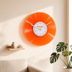 Op de afbeelding: Een ronde wandklok gemaakt van een doorschijnende oranje vinylplaat. Het midden heeft een witte wijzerplaat met oranje cijfers en de tekst "Midnights" in het midden gedrukt. De klok heeft twee dunne witte wijzers die de tijd aangeven.
