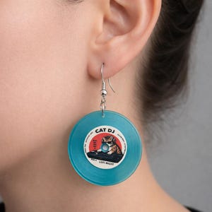 Könnte beinhalten: Türkisfarbene Vinyl-Schallplatten-Ohrringe mit einem silbernen Haken. Das mittlere Etikett zeigt eine Katze mit Sonnenbrille und Kopfhörern, mit dem Text "CAT DJ" und "LOFI NIGHT". Ein einzigartiges Accessoire.