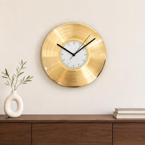 Könnte beinhalten: Eine runde Wanduhr, die wie eine goldene Schallplatte gestaltet ist. Die Uhr verfügt über ein weißes Zifferblatt in der Mitte mit schwarzen Ziffern von 1 bis 12 sowie dünnen schwarzen Zeigern für Stunden, Minuten und Sekunden.