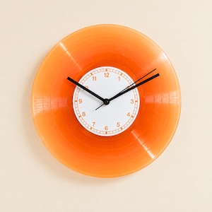 Op de afbeelding: Een ronde wandklok gemaakt van een doorschijnende oranje vinylplaat. Het midden heeft een witte wijzerplaat met oranje cijfers van 1 tot 12 en een rasterachtige rand. Drie dunne zwarte wijzers geven de tijd aan op de wijzerplaat.