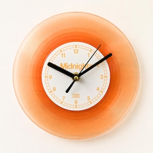 Peut inclure: Une horloge murale circulaire fabriquée à partir d'un disque vinyle orange translucide. Le centre présente un cadran blanc avec des chiffres orange de 1 à 12, le mot Midnights et le logo Republic. Trois aiguilles noires sont au centre.