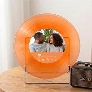 Op de afbeelding: Oranje vinylplaat met een foto van een stel in het midden. De plaat wordt op een metalen standaard gepresenteerd. Het heeft een muziekspelerinterface met het woord "Swift" en de songtitel "Lover".
