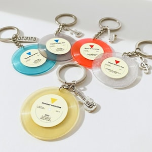 May include: A collection of colorful vinyl record keychains. Each keychain features a miniature record with text, a metal charm, and a keyring. The records are in various colors, including blue, red, yellow, and clear.