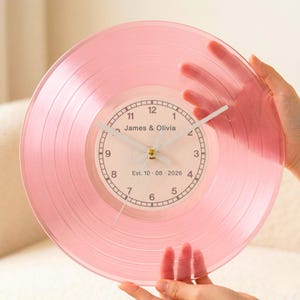 May include: A pink, transparent vinyl record clock. The clock face features the names "James & Olivia" and the date "Est. 10.08.2026". The clock has white hands and a gold-colored center.