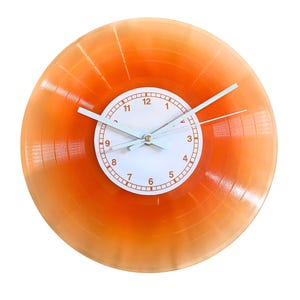 May include: A circular wall clock featuring a translucent orange vinyl record as the base. The centre has a white circular face with orange numerals from 1 to 12 and thin white hour, minute, and second hands attached to a central gold pin.