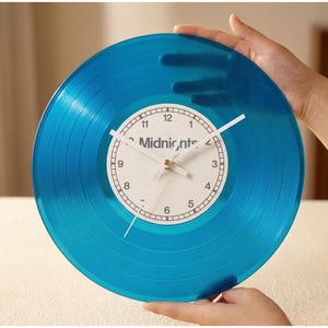 May include: A blue vinyl record clock with white clock hands and a white clock face. The clock face has the word "Midnights" printed on it. The clock is held by two hands.