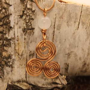 May include: A copper pendant necklace with a three-spiral design. The pendant hangs from a fine gold-coloured chain and has a white, round bead at the top. The necklace is set against a textured, light-coloured background.