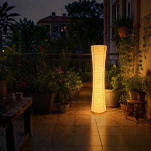 May include: A tall, cylindrical floor lamp with a textured, off-white paper shade that emits a warm, golden glow. The lamp features a slender, slightly tapered waist design and stands upright on a flat base, illuminating the surrounding area.