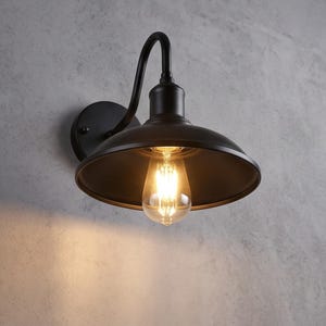 Op de afbeelding: Een matzwarte wandlamp met een gebogen arm en een brede, komvormige kap. Het armatuur bevat een enkele, zichtbare Edison-stijl gloeilamp die een warme, gouden gloed uitstraalt en het gebied direct onder de lampenkap verlicht.