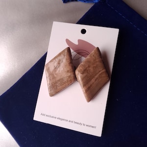 May include: A pair of brown, diamond-shaped earrings with a marbled appearance, displayed on a white card. The card is placed on a navy blue velvet pouch. Text on the card reads: "Add exclusive elegance and beauty to women!"