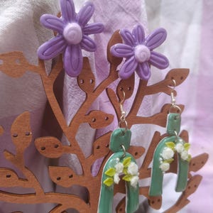 May include: A pair of earrings and two flower-shaped stud earrings. The earrings are green with white floral accents. The stud earrings are purple with a white center. The earrings are displayed on a wooden tree-shaped stand.