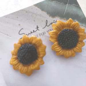 Puede incluir: Un par de pendientes en forma de girasol con pétalos amarillos y un centro negro texturizado, sujetos a una tarjeta expositora blanca con el texto Sweet lovin' y JUST FOR YOU en letras negras cursivas y de molde.