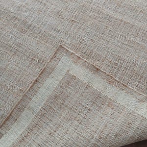 Peut inclure: Gros plan sur un tapis beige tiss&eacute; avec une surface textur&eacute;e. Le tapis a une teinte naturelle et terreuse avec des fibres visibles et un motif subtil. Le coin du tapis est repli&eacute;, r&eacute;v&eacute;lant une bordure plus claire.